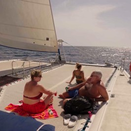 Three people relaxing on a sailboat deck under bright sunlight, with ocean in the background.