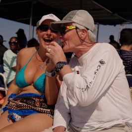 Two people laughing and talking on a boat with others in the background.