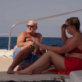 Two people relaxing on a yacht deck with drinks, ocean in background.