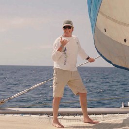 Person on a boat holding a sail, wearing sunglasses and casual attire, with the ocean in the background.