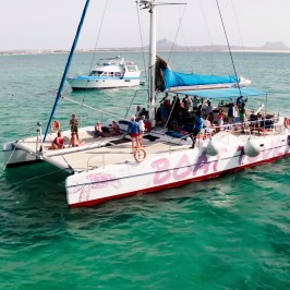 A catamaran with people on board sails in clear turquoise water, with another boat in the background.