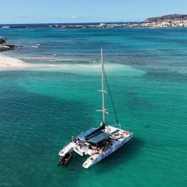 Catamaran with people on turquoise water near a sandy beach and distant town.