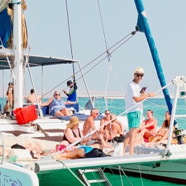 People sunbathing on a catamaran, with some swimming in the turquoise sea.